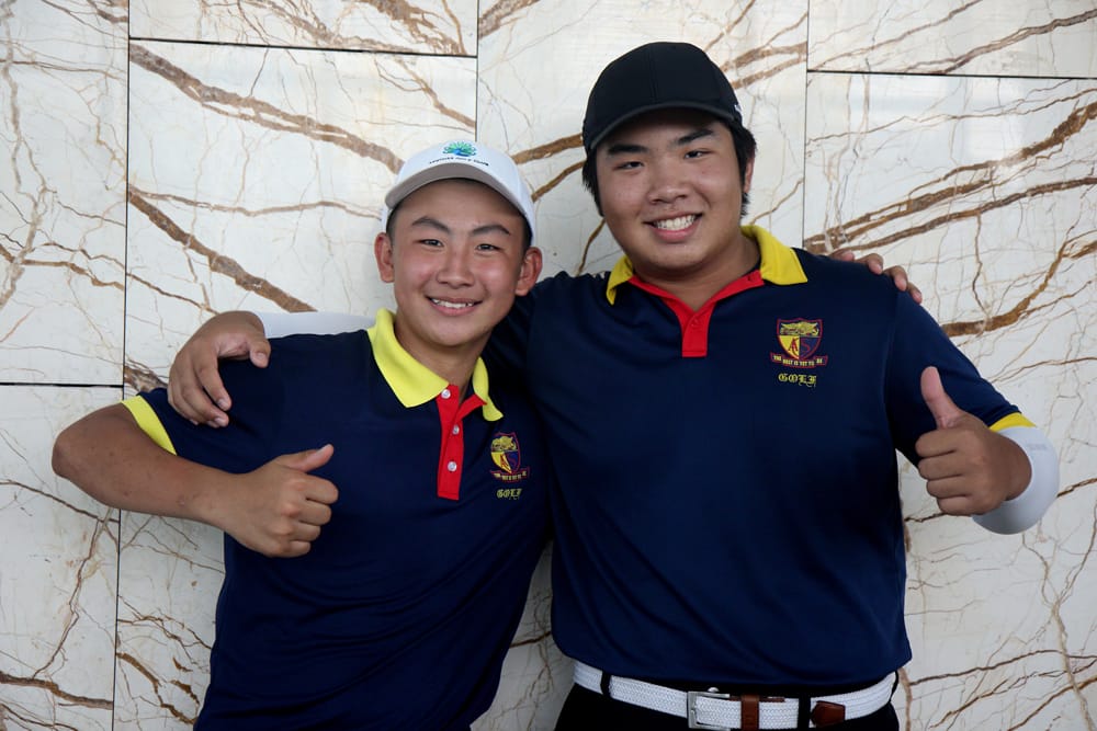 Two smiling young men in matching navy and yellow golf polo shirts giving thumbs up, posed in front of a marble wall.